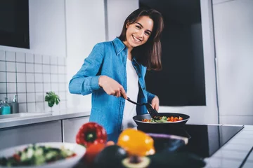 Abwaschbare Fototapete Kochen Glücklich lächelnde Frau, die in der Küche zu Hause frisches Gemüse kocht, glückliches brünettes Mädchen, das veganes Essen auf Elektroherd in der modernen Küche kocht, gesunder Ernährungslebensstil