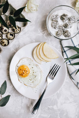Breakfast of fried egg and pear on a white plate with a fork. In a glass bowl, yogurt and a dragon fruit, next to a tray of quail eggs, tablecloth and flowers on a gray concrete background flatlay