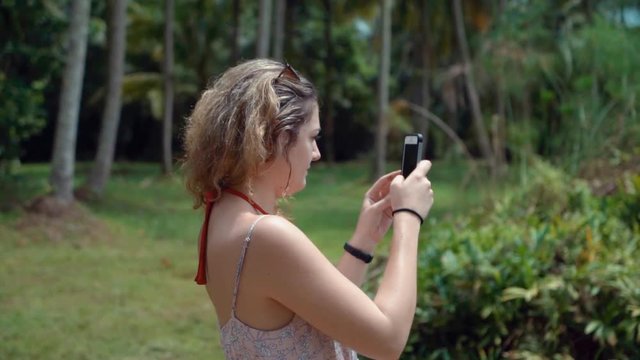 Beautiful young blonde millennial woman takes a panoramic video with her smartphone in a forest in Singapore in slow motion
