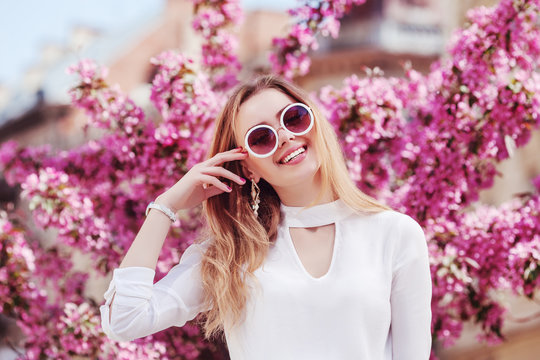 Outdoor portrait of young beautiful happy smiling girl wearing stylish round sunglasses and white blouse, posing in street with spring blooming pink trees. Female fashion, beuty concept