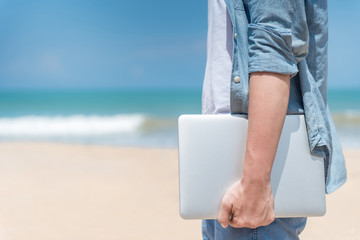 Male hand holding laptop on the beach, working outdoor in summer season, digital nomad man...