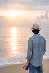 Young Asian man traveler and photographer with jean shirt and hat holding film camera and looking beautiful sunset at tropical beach island. Summer holiday or vacation travel concepts