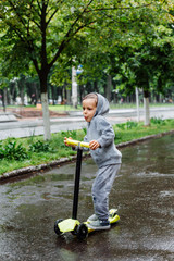 Drenched in the rain, a boy in a sport suit skates on a scooter. Spring walk in the city park, rainy weather.