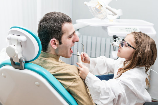 Little girl in a white robe in the image of a dentist examines teeth to a man sitting in a dental chair