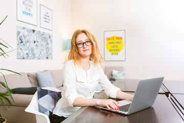 Young woman with long red hair and glasses for view works, prints on a gray laptop keyboard sitting...