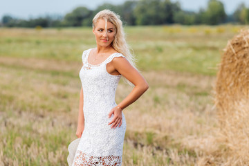 Fototapeta na wymiar Beautiful tanned girl in white lace dress is standing near haystack at sunset in the summer. Happy, smiling woman in straw hat having fun, posing at the field. Concept of countryside lifestyle.