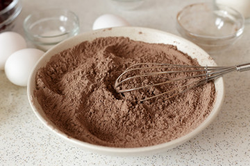 Mixture of flour and cocoa powder in bowl with the whisk on the kitchen table.
