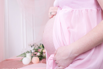 Close up of pregnant woman touching her belly. Woman body during pregnancy in pink dress on background with flower. Her hands on stomach, hands over tummy. Expectation conсept