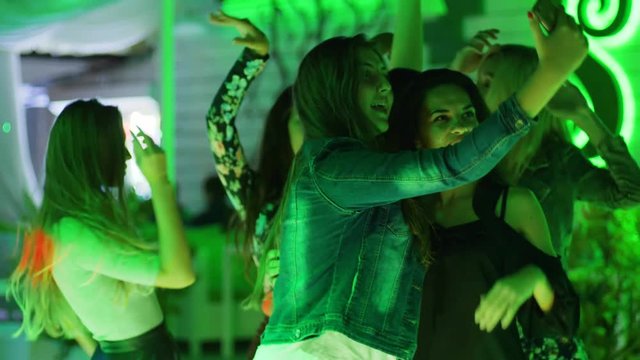 Funny girls have fun and take photos on gadget on background of dancing people at nightclub hen-party,