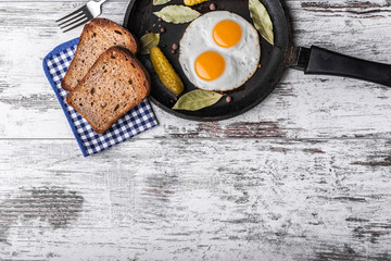 Fried eggs in a frying pan. Rye toast and pickled cucumber next to fried eggs.