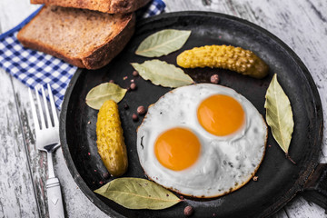 Fried eggs in a frying pan. Rye toast and pickled cucumber next to fried eggs.