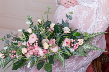 Close up of pregnant woman touching her belly in white lace dress holding green and pink bouquet. Motherhood, pregnancy, expectation and spring concept. Pregnant woman expecting baby.