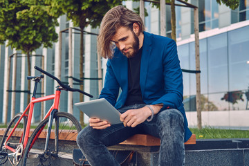 Fototapeta na wymiar A handsome fashionable bearded male with long hair sitting on a bench with a bicycle, using a tablet computer.