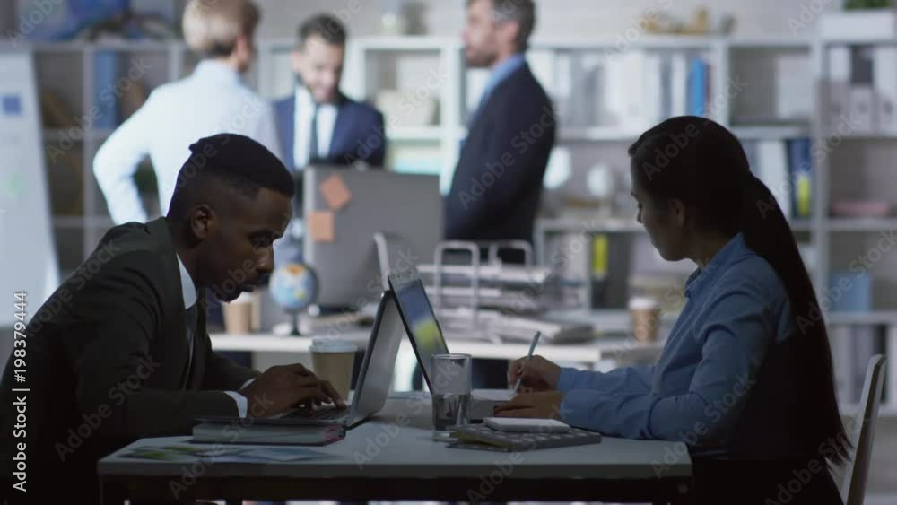 Poster side view of two office workers sitting at desk opposite each other and using laptop computers, thei