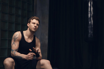 Indoor shot of thoughtful attractive young European man wearing sleeveless shirt and shorts wrapping boxing bandages, sitting against black curtain background with copyspace for your information
