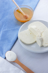 Cheese, honey and sour cream, feta in a white plate, soft cheese on a white background, wooden spoon with sour cream, French breakfast, blue napkin, honey in glassware, art