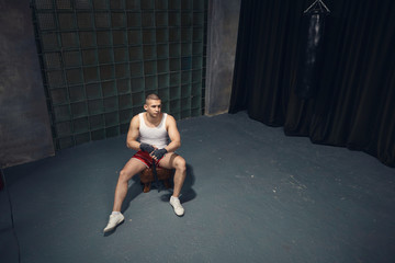 Fototapeta na wymiar Top view of fashionable serious young muscular businessman wearing white sleeveless shirt, sneakers and red trousers taping hands with bandages before boxing training after working day at office