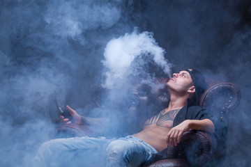 Vaper. The man with tattoos sits on a leather sofa smoke an electronic cigarette on the dark...