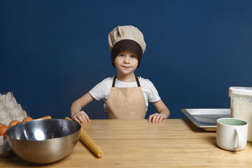 Picture of determined excited little boy wearing chef uniform standing at kitchen table with metal bowl, rolling pin, tray, flour and eggs, ready to start making dough for pancakes. Food and nutrition