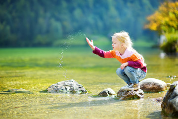 Adorable girl playing by Konigssee lake in Germany on warm summer day. Cute child having fun feeding ducks and throwing stones into the lake. - obrazy, fototapety, plakaty