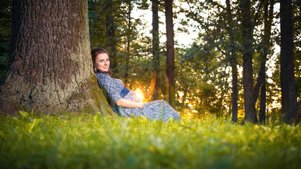 Happy pregnant young woman sitting leaning against a huge tree