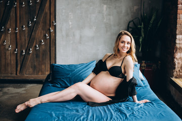 Beautiful pregnant woman in black lingerie with charming smile lies on the bed in the bedroom
