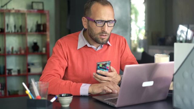 Young man working with smartphone and laptop sitting by table at home

