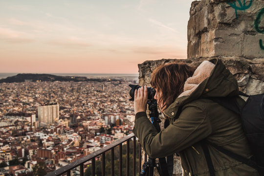 A woman photographer is taking a picture to the views of a big city with a camera and a tripod. She wears a green coat with hood.
