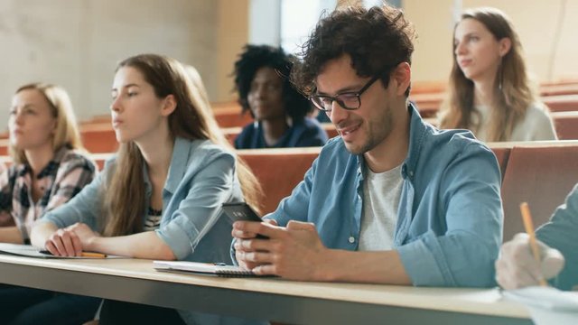 Hispanic Man Uses Smartphone While Being on a Lecture in the Classroom. Lecture Hall Filled with Students Studying. Young People at University. Shot on RED EPIC-W 8K Helium Cinema Camera.