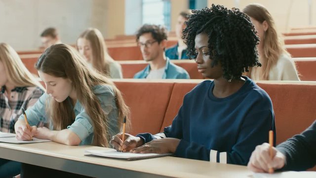 Beautiful Black Female Student Sitting Among Her Fellow Students in the Classroom, She's Writing in the Notebook and Listens to a Lecture. Shot on RED EPIC-W 8K Helium Cinema Camera.