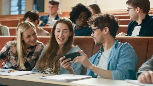 Male Student Shares Mobile Phone Screen with Fellow Students, they Smile. Joking in the University Classroom. Shot on RED EPIC-W 8K Helium Cinema Camera.