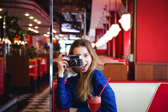 Indoor fashion portrait of pretty young brunette girl sitting at big white bed and taking picture on vintage retro old school camera, wearing cute home outfit, having fun alone.