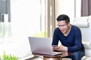 Happy excited asian man celebrate with smartphone and computer laptop, success or happy action. Freelancer or Entrepreneur using technology and start up modern office with copy space