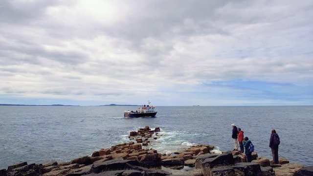 People are staying on volcanic rocks and watching a sailing boat. Group of tourists looking at ship passing by close to the shore of Staffa Island, near famous Fingal's Cave with basalt columns.