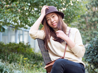 Outdoor portrait of young beautiful fashionable playful Chinese lady sitting and posing on bench. Model wearing stylish hat & clothes. Sunny day. Female fashion. City lifestyle.