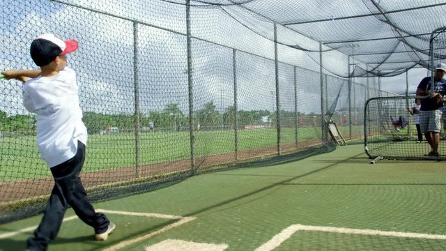 Slow motion shot of a man throwing ball to kid inside batting cages at baseball park. Kid misses.