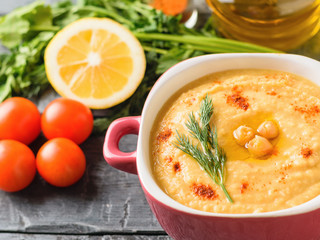 Fresh hummus in a purple bowl, tomatoes, lemon and a bunch of greenery on a rustic table.