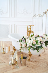 Beautiful luxury vintage empty bathtub with lush floral decorations and candles, mirror on the floor in bathroom interio, copy space. Freestanding white bath with flowers near folding screen