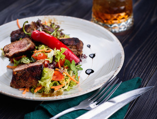 Veal salad with lettuce, carrot, tomato and pepper on white plate on dark wooden background, copy space. Restaurant food, close up. Meat salad and glass of whiskey