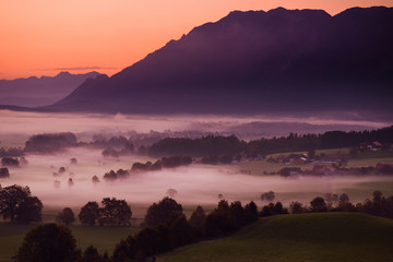Obraz premium Breathtaking morning lansdcape of small bavarian village covered in fog. Scenic view of Bavarian Alps at sunrise with majestic mountains in the background.