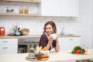 A young girl in a bathrobe with a cup of tea in her hands is sitting at a kitchen table smiling and looking away. Home style. Lifestyle. Space for text