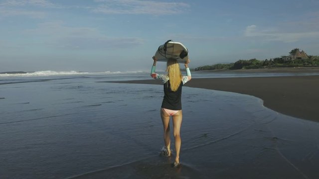 Young woman surfer walking at sandy beach against blue water and holding surfboard during the sunrise
