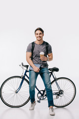 Fototapeta na wymiar smiling student holding coffee to go and leaning on bicycle on white
