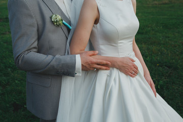 Elegant wedding couple is hugging outdoors. Groom in checkered grey suit and tiffany light blue bow tie. Bride in satin white dress. Summer romance. wedding buttonhole and white golden rings.