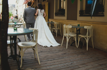 Elegant wedding couple is walking in old ancient town street cafe. Slim bride in satin dress with train. Groom in grey checkered suit. Wooden floor and summer atmosphere.