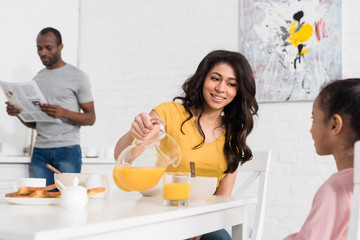 mother pouring orange juice for daughter on breakfast while father reading newspaper blurred on background