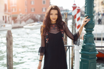 Naklejka premium Attractive young romantic woman standing on the pier against beautiful view on venetian chanal with boats and gondolas in Venice, Italy. Travel tourist girl on vacation walking happy by Grand Canal