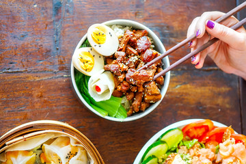 Braised pork belly rice bowl on a wooden table with egg and salad. A hand with chopsticks holds a piece of meat. Top view with copy space