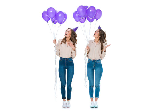 surprised attractive young twins looking at violet balloons isolated on white