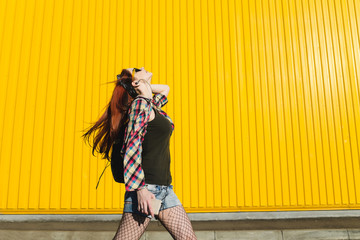 Redhead girl in denim shorts and pantyhose mesh is listening music with headphones on yellow background